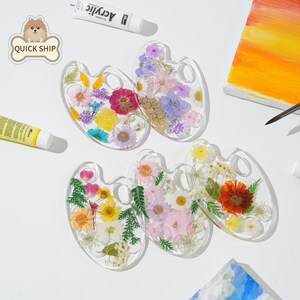 May include: Five clear resin palettes shaped like artist palettes, each embedded with a variety of colorful pressed flowers and green foliage. The palettes are arranged on a white surface alongside a tube of acrylic paint and a canvas.