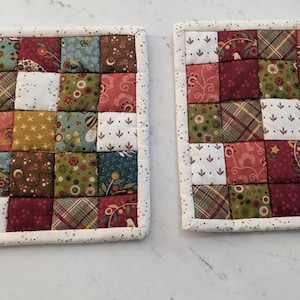 May include: Two square quilted pot holders. Each pot holder is made of small fabric squares in various colors and patterns, including floral, plaid, and solid colors. The edges are finished with a dotted white fabric.