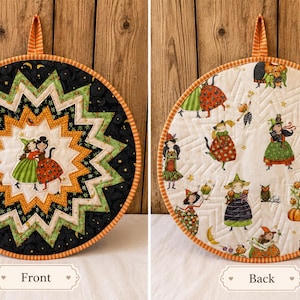 Peut inclure: Un dessous de plat matelassé rond avec un motif en étoile aux tissus noirs, verts et orange sur le devant, et un fond blanc avec un motif répétitif de sorcières, citrouilles et hiboux au dos, fini par une boucle de suspension orange.