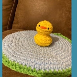May include: A handcrafted yellow crochet duck with black eyes and an orange beak sits on a light blue and green crochet pad. The duck is the main focus, highlighting the item's craftsmanship.