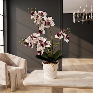 Op de afbeelding: Een kunstmatige orchidee met witte bloemblaadjes met donkerpaarse stippen en dieppaarse harten. De plant heeft groene bladeren en staat in een witte, cilindrische pot met een geweven patroon op het buitenoppervlak van de pot.