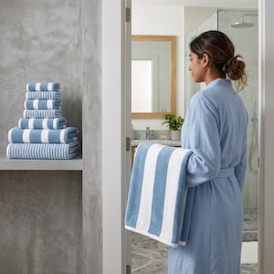 Pode incluir: Uma pilha de toalhas às riscas azuis e brancas de vários tamanhos sobre uma prateleira. Veste-se um roupão azul claro e segura-se uma toalha a condizer às riscas azuis e brancas. As toalhas apresentam riscas verticais grossas e alternadas.