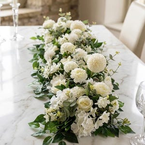 May include: A long floral table runner featuring an arrangement of cream and white artificial flowers, including round pom-pom blooms, roses, and hydrangeas, interspersed with green eucalyptus leaves and various foliage on a white marble surface.