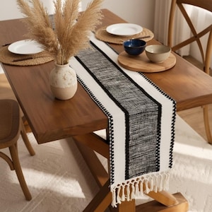 Könnte beinhalten: Ein Holztisch ist mit einem schwarz-weiß gewebten Tischläufer mit Fransen gedeckt. Eine Vase mit getrocknetem Pampasgras steht auf dem Tisch, zusammen mit Tellern und Schalen. Der Tisch ist aus Holz.