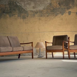 May include: A mid-century modern furniture set featuring a brown two-seater sofa and two armchairs with wooden frames and brown cushions. A small table lamp with a beige shade sits between the chairs. The setting is a room with a textured wall.