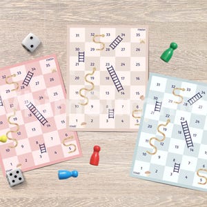 May include: Three colorful Snakes and Ladders board games with dice and game pieces. The boards are pink, beige, and blue, each with a grid of numbers, ladders, and snakes. The game pieces are yellow, red, blue, and green.