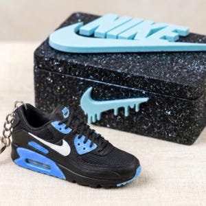 Puede incluir: Un llavero de zapatilla negro, blanco y azul claro con un zapato deportivo en miniatura detallado y una anilla de metal. Detrás hay una caja rectangular negra con acabado brillante, adornada con el logotipo y el texto de Nike en azul claro.