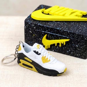 K&ouml;nnte beinhalten: Ein Miniatur-Schl&uuml;sselanh&auml;nger mit wei&szlig;en, schwarzen und gelben Turnschuhen mit dem Nike Air Max-Logo. Daneben eine schwarze Box mit gelbem Nike-Logo. Die Box hat eine glitzernde Textur.