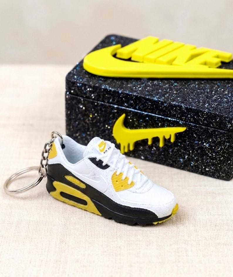 K&ouml;nnte beinhalten: Ein Miniatur-Schl&uuml;sselanh&auml;nger mit wei&szlig;en, schwarzen und gelben Turnschuhen mit dem Nike Air Max-Logo. Daneben eine schwarze Box mit gelbem Nike-Logo. Die Box hat eine glitzernde Textur.