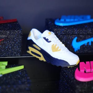 K&ouml;nnte beinhalten: Wei&szlig;er, marineblauer und gelber Sneaker mit sichtbarer Air-Unit. Der Schuh hat ein klassisches Design mit dem Nike-Logo. Weitere Sneaker mit unterschiedlichen Farbschemata sind im Hintergrund zu sehen.