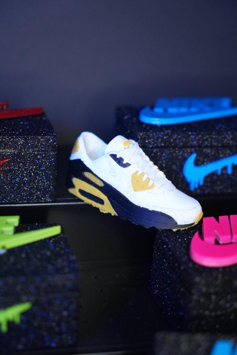 K&ouml;nnte beinhalten: Wei&szlig;er, marineblauer und gelber Sneaker mit sichtbarer Air-Unit. Der Schuh hat ein klassisches Design mit dem Nike-Logo. Weitere Sneaker mit unterschiedlichen Farbschemata sind im Hintergrund zu sehen.