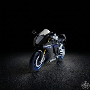 May include: A black and blue Yamaha R1 motorcycle model. The sport bike has a white windscreen, blue accents on the wheels and body, and the text "R1" in blue. The motorcycle is on a dark asphalt surface.