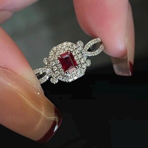 Pode incluir: Um anel de prata com uma pedra preciosa retangular, vermelho escuro no centro, rodeada por pedras menores e transparentes. A banda do anel apresenta um design torcido, e a cravação é intrincada e detalhada.