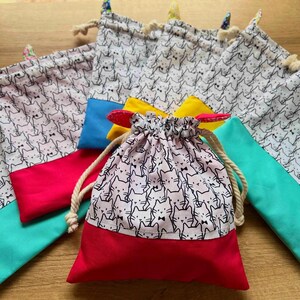 May include: A collection of drawstring bags with a repeating cat face pattern. The bags have a white background with black cat faces and are trimmed with red, teal, yellow, and blue fabric. The bags are tied with a natural rope.