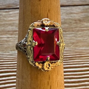 Pode incluir: Um anel com uma grande pedra retangular facetada de cor vermelho-profundo, montada numa armação de metal decorativa. O engaste apresenta um trabalho de filigrana intrincado nas laterais e detalhes florais dourados no topo e na base.