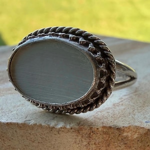 May include: A silver ring featuring an oval-shaped, pale gray, smooth stone set within a twisted rope-style metal border. The band of the ring is split into two separate silver wires that connect to the base of the oval setting on each side.