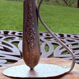 May include: A silver-toned metal bud vase crafted from a repurposed spoon. The vertical handle features an embossed floral pattern, while the bowl of the spoon is flattened to form a circular base. A curved metal arm extends from the base to the top.