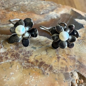 May include: A pair of silver-toned flower earrings with a pearl center. The earrings have dark gray petals and are set against a light brown and tan stone surface. The earrings are designed to be worn on the ear.