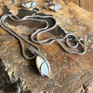 May include: A silver necklace with a pendant and matching earrings. The pendant and earrings feature a white, teardrop-shaped stone set in a silver frame. The necklace chain is a fine, textured silver.