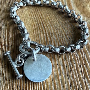May include: A silver-toned chain link bracelet featuring a toggle clasp and a circular pendant. The bracelet is constructed from rounded, interconnected metal links, with a smooth, flat, round disc charm attached to the toggle ring mechanism.