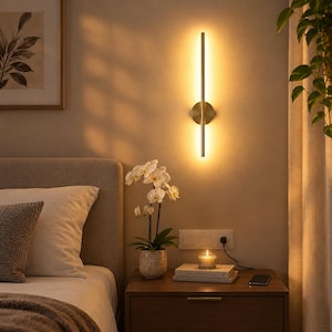 May include: A vertical wall-mounted light fixture with a slim, cylindrical gold-toned metal bar and a circular base plate. The lamp emits a warm, diffused glow against the wall, creating a soft illumination effect in the room.