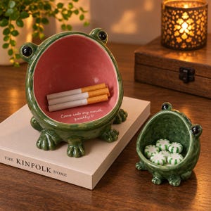 May include: Two green ceramic frog-shaped bowls with black eyes and pink interiors. The larger bowl contains three cigarettes and features the white text "Come into my mouth quickly!" with a heart. The smaller bowl holds several round white and green candies.