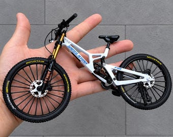 May include: A miniature white mountain bike model featuring black tyres with yellow lettering, gold-coloured front suspension forks, and a black seat. The frame displays the text "SHILILAIZAD" and "FSR" in blue, with black spokes and disc brakes.