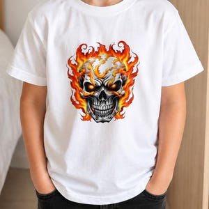 May include: A white short-sleeved t-shirt featuring a central graphic print of a human skull engulfed in vibrant orange and yellow flames. The skull has glowing orange eyes and detailed shading, creating a high-contrast design on the plain fabric.