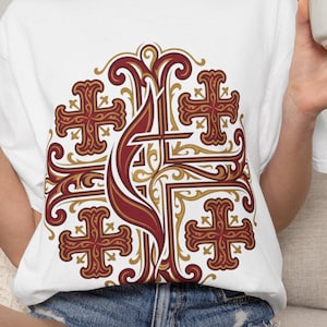 May include: A white short-sleeved t-shirt featuring a large, intricate graphic design on the chest. The design displays a central cross motif in shades of deep red and gold, surrounded by four smaller ornate crosses in a symmetrical pattern.