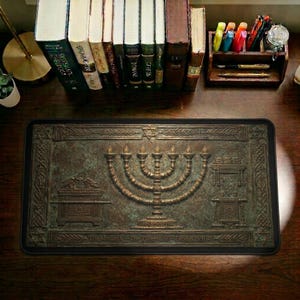 Pode incluir: Tapete de mesa retangular bronzeado com um relevo detalhado de um menorá, a Arca da Aliança e uma mesa. O texto "The Menorah - The Tabernacle - Exodus 25:31-40" está na parte inferior. O tapete está em uma mesa de madeira com livros e acessórios de mesa.