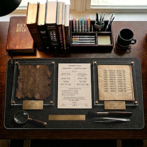 May include: A desk setup featuring books, pens, and historical documents. The display includes a Bible, a wooden pen holder, and framed ancient texts. A magnifying glass and other tools are also present, suggesting a study or research environment.
