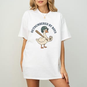 May include: White t-shirt featuring a cartoon goose wearing a baseball helmet and holding a bat and coffee cup. The shirt has the text "OUTNUMBERED BY BOYS" in a circular design around the goose.