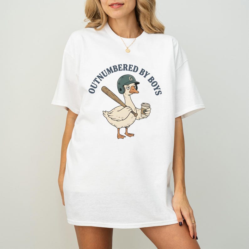 May include: White t-shirt featuring a cartoon goose wearing a baseball helmet and holding a bat and coffee cup. The shirt has the text "OUTNUMBERED BY BOYS" in a circular design around the goose.