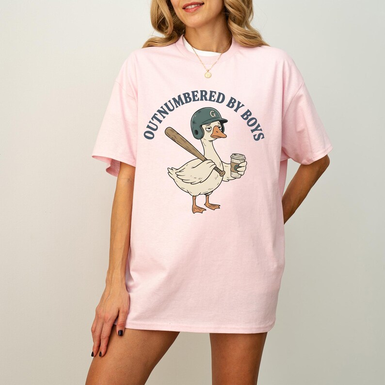 May include: Pink t-shirt with the text "OUTNUMBERED BY BOYS" in a circular design. The shirt features a cartoon goose wearing a baseball helmet and holding a bat and coffee cup. The goose is light beige with brown and orange accents.