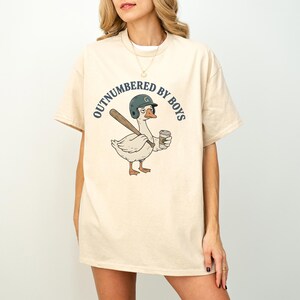 May include: Cream-colored t-shirt featuring a cartoon goose wearing a baseball helmet and holding a bat and coffee cup. The text "OUTNUMBERED BY BOYS" is arched above the goose. The shirt has short sleeves and a relaxed fit.