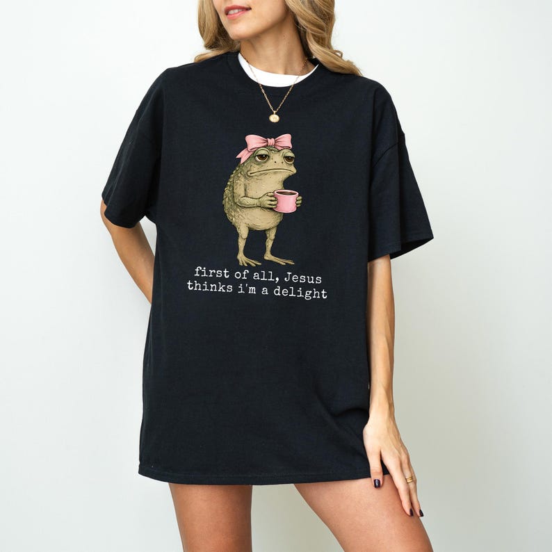 Peut inclure: T-shirt noir avec une illustration fantaisiste d'une grenouille portant un n&oelig;ud rose et tenant une tasse rose. Le texte "first of all, Jesus thinks I'm a delight" est imprim&eacute; en dessous.