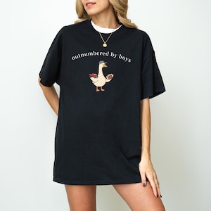 May include: Black t-shirt with the phrase "outnumbered by boys" in white script. The shirt features a cartoon goose wearing a hat and holding a football. The goose has a red and white floral design on its back.