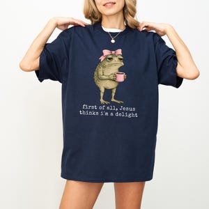 Peut inclure: T-shirt bleu marine avec une grenouille de dessin anim&eacute; portant un n&oelig;ud rose et tenant une tasse rose. Le texte "first of all, Jesus thinks I'm a delight" est imprim&eacute; en dessous.