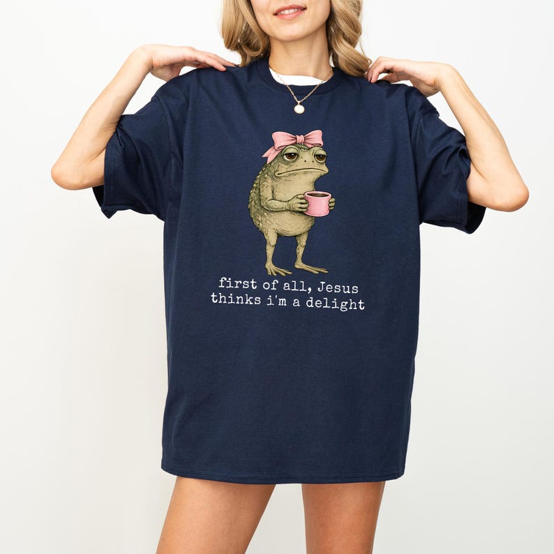 Peut inclure: T-shirt bleu marine avec une grenouille de dessin anim&eacute; portant un n&oelig;ud rose et tenant une tasse rose. Le texte "first of all, Jesus thinks I'm a delight" est imprim&eacute; en dessous.