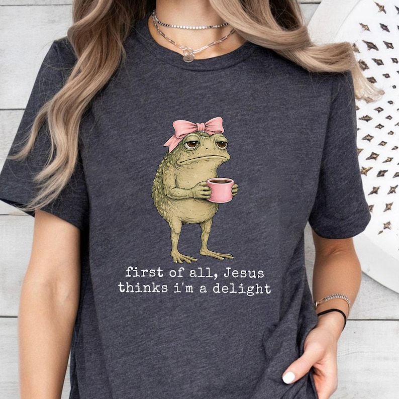 Peut inclure: T-shirt gris fonc&eacute; avec une grenouille de dessin anim&eacute; portant un n&oelig;ud rose et tenant une tasse rose. Le texte sur le t-shirt se lit comme suit : "first of all, Jesus thinks I'm a delight."