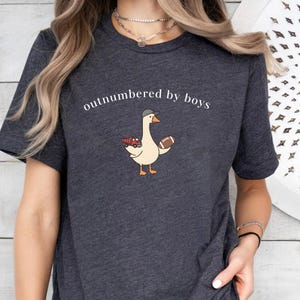 May include: Dark gray t-shirt with the text "outnumbered by boys." The shirt features a cartoon goose wearing a baseball cap and holding a football and a red truck. The goose is light beige with orange feet and beak.