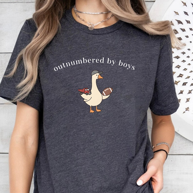 May include: Dark gray t-shirt with the text "outnumbered by boys." The shirt features a cartoon goose wearing a baseball cap and holding a football and a red truck. The goose is light beige with orange feet and beak.