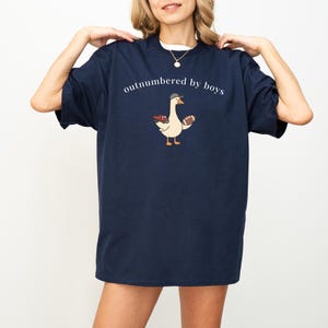 May include: Navy blue t-shirt with the phrase "outnumbered by boys" in white. A cartoon goose wearing a cap and holding a football is printed below the text. The goose also has a small red truck.