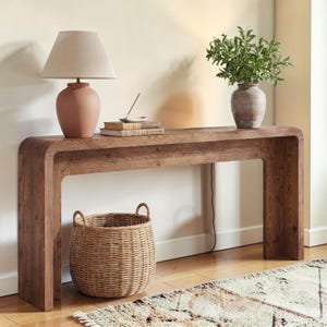 Puede incluir: Una consola de madera con un diseño curvo, que sostiene una lámpara de terracota, libros y un jarrón con vegetación. Una cesta tejida se encuentra debajo. La mesa es de madera marrón claro, y la lámpara tiene una pantalla beige.