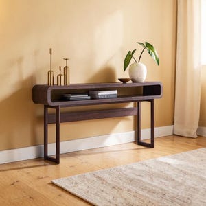 May include: A dark brown, modern console table with a curved top and open shelf. The table has a rectangular base and holds decorative items, including gold candlesticks, books, and a vase with greenery. The table is set against a yellow wall.