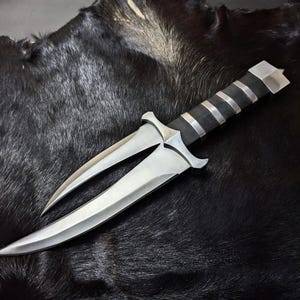 May include: A double-bladed dagger featuring two curved, silver-toned metal blades that extend from a central guard. The handle is black with three silver-toned metal bands and a flat, silver-toned pommel at the base of the grip.