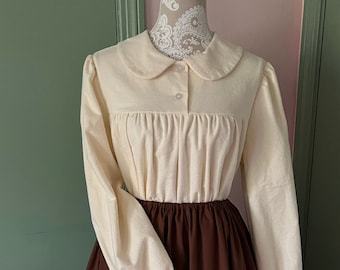May include: A white long-sleeved blouse with a peter pan collar and a brown skirt. The blouse has a button closure and a gathered bodice.