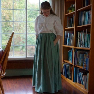 Könnte beinhalten: Eine Frau in einem weißen Oberteil und einem langen, salbeigrünen Rock steht in einem Raum mit einem Bücherregal aus Holz, das mit Büchern gefüllt ist, einem Tisch mit Stühlen und einem Fenster mit Blick auf Bäume.