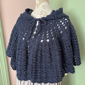 Op de afbeelding: Een marineblauwe, gehaakte cape met een capuchon en striksluiting. De cape heeft een geschulpte rand en een openwerk ontwerp. Het kledingstuk wordt op een mannequin getoond.