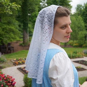 May include: A woman wearing a white lace head covering and a light blue dress stands in a garden setting.